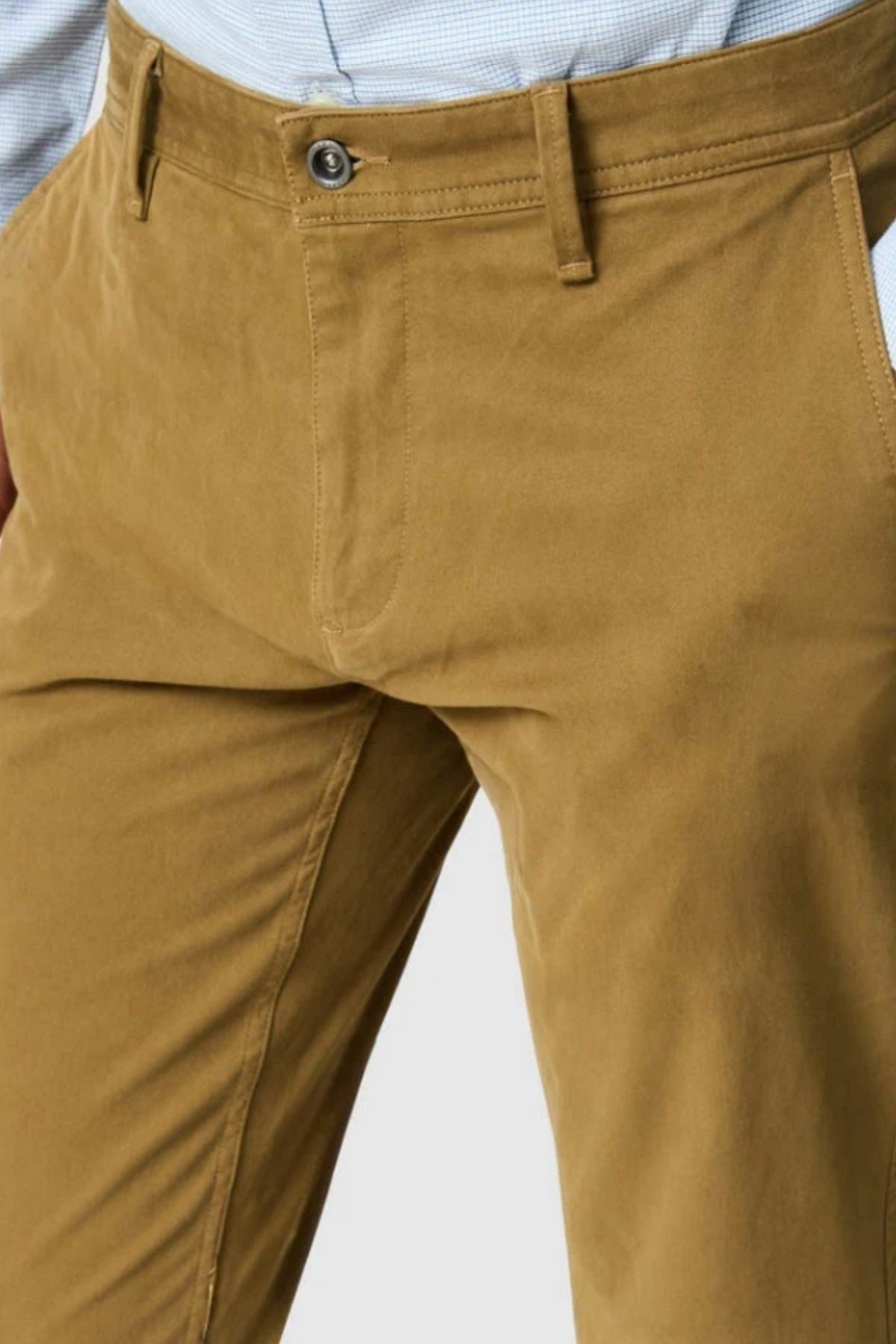 Close-up of a person wearing Rodd & Gunn Thomas Road 2.0 Chino Pant in tan, paired with a light blue checkered shirt tucked in. Only the waist and upper legs are visible; hands rest casually in their pockets.