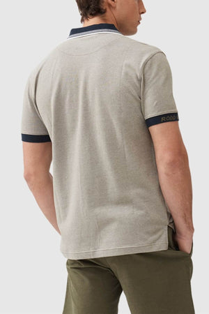 A person confidently stands with their hands in olive pants' pockets, wearing a Rodd & Gunn Masterton Sports Fit Polo made from premium USA cotton. The polo features dark trim on the collar and sleeves, and "RODD" is visible on the sleeve against a plain white background.