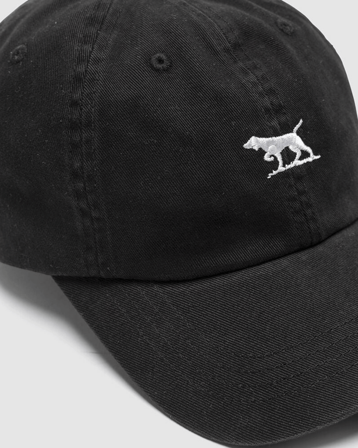 The Rodd & Gunn Gunn Cap is a black cotton twill baseball cap with a small white embroidered dog logo on the front. It features an adjustable strap for a perfect fit and is pictured at an angle against a light gray background.