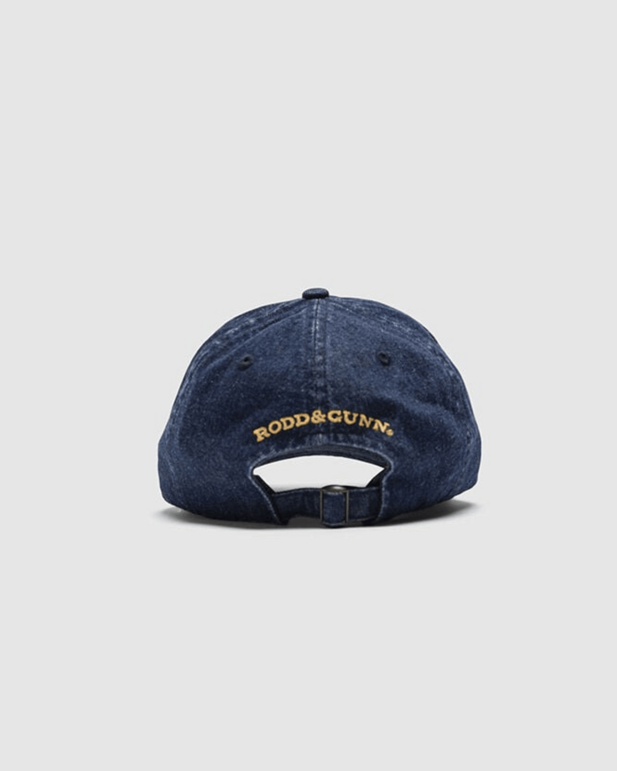 The Rodd & Gunn Gunn Cap in dark blue heavy-duty cotton twill is shown from the back, with a brass buckle strap and yellow "RODD & GUNN." embroidery above the opening.