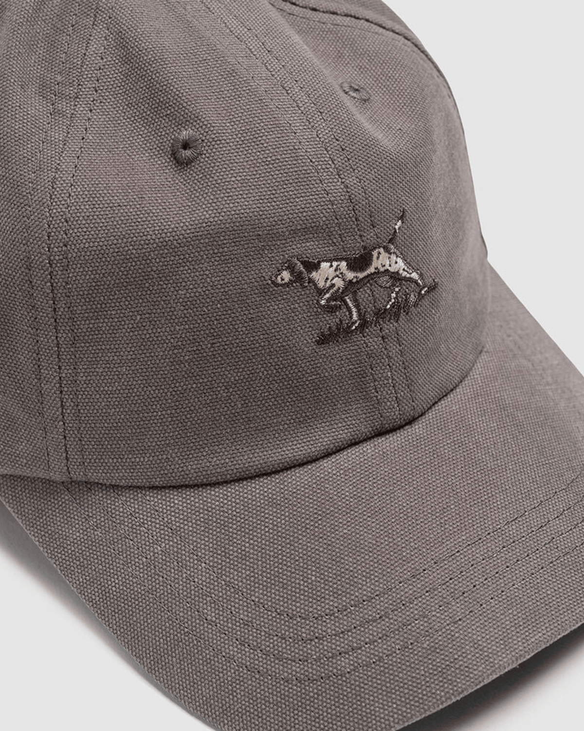 A close-up of the Rodd & Gunn Signature Cap, a gray cotton twill cap with an embroidered black and white dachshund on the front, visible stitching, metal eyelets, and classic brand style.