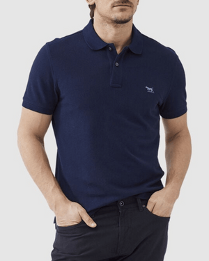 A man is wearing a Rodd & Gunn The Gunn Polo in navy blue, featuring a modern fit made from Australian cotton and adorned with a small animal logo on the left chest. He pairs it with dark pants and has his hands in his pockets, standing against a plain light background.