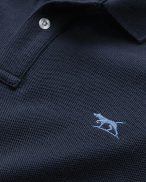 Close-up of the Rodd & Gunn The Gunn Polo in navy blue, showcasing a light blue embroidered dog logo on the chest. The comfortable shirt is made from premium Australian cotton with a textured fabric, featuring a modern fit and two buttons visible near the collar.