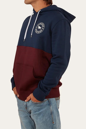 Someone is wearing the Ringers Western Hahndorf Hoodie, featuring a navy top, maroon bottom, white drawstrings, kangaroo pocket, and circular chest logo in brushed fleece, paired with blue jeans.