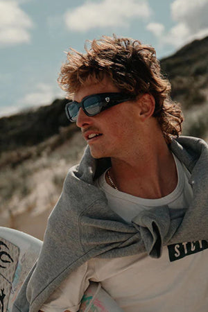 A man with a mullet and sunglasses, wearing a white t-shirt and gray sweatshirt over his shoulders, stands outdoors holding a surfboard. He wears SafeStyle Auras Black Frame Grey Polarised Lens, AS/NZS 1337.1 compliant for eye protection.