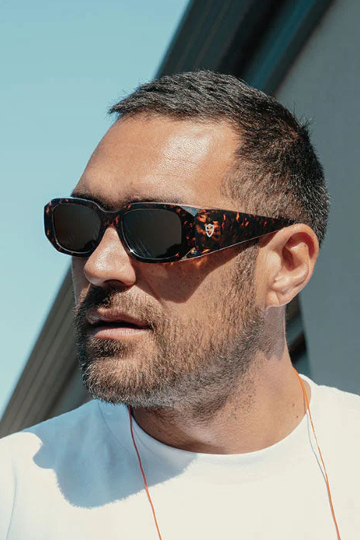 A man with short dark hair and a beard wears SafeStyle Auras Brown Tort Frame Brown Polarised Lens sunglasses and a white t-shirt, looking to the side outdoors with a building and blue sky in the background.