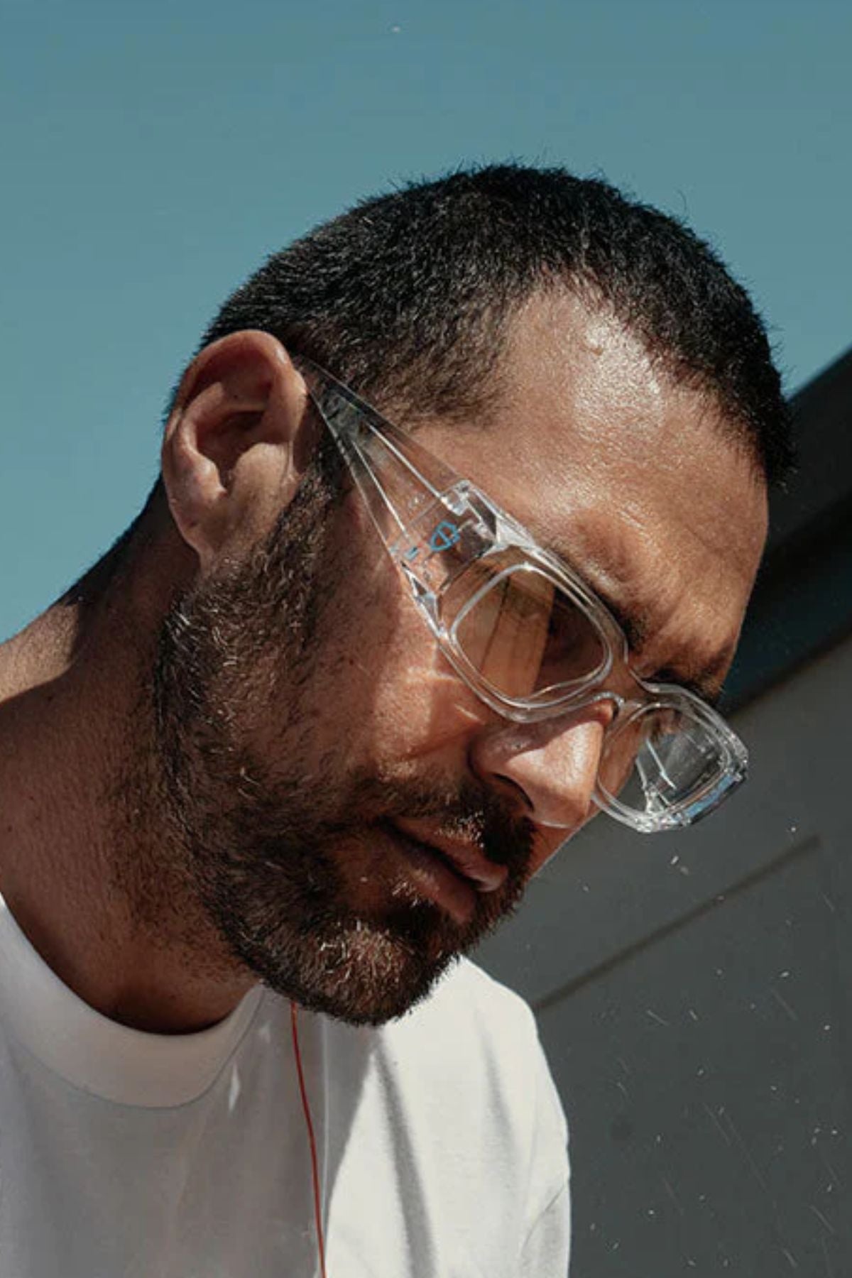 A man with a short beard and close-cropped hair wears SafeStyle Auras Clear Frame Clear Lens eye protection and a white shirt, looking downward outdoors under a blue sky.