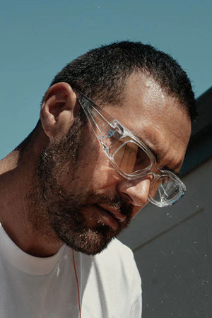 A man with a short beard and close-cropped hair wears SafeStyle Auras Clear Frame Clear Lens eye protection and a white shirt, looking downward outdoors under a blue sky.
