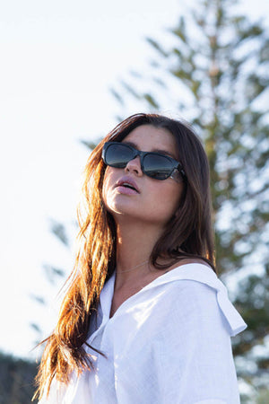 A woman with long brown hair wears SafeStyle Classics Black Frame sunglasses with polarised lenses and UV protection, gazing upwards against a backdrop of trees and a clear sky.