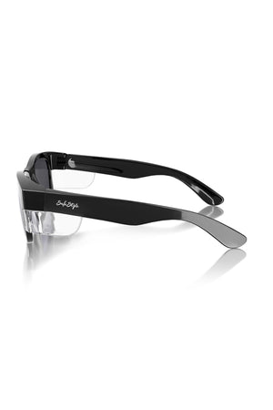 Side view of SafeStyle Classics Black Frame Polarised Lens sunglasses with clear UV-protection lenses, featuring thick temples and a reflective silver logo on one arm. The sleek black glasses rest atop a white reflective surface.