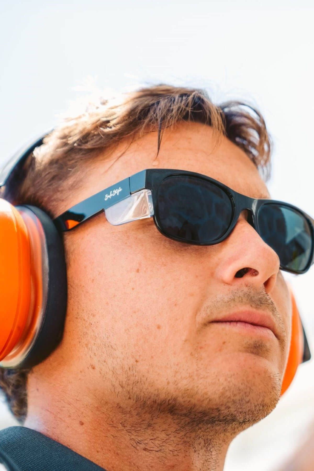A person with short hair is wearing large orange earmuffs and SafeStyle Classics Black Frame Tinted Lenses sunglasses, providing UV protection. The person appears to be looking into the distance against a light background.