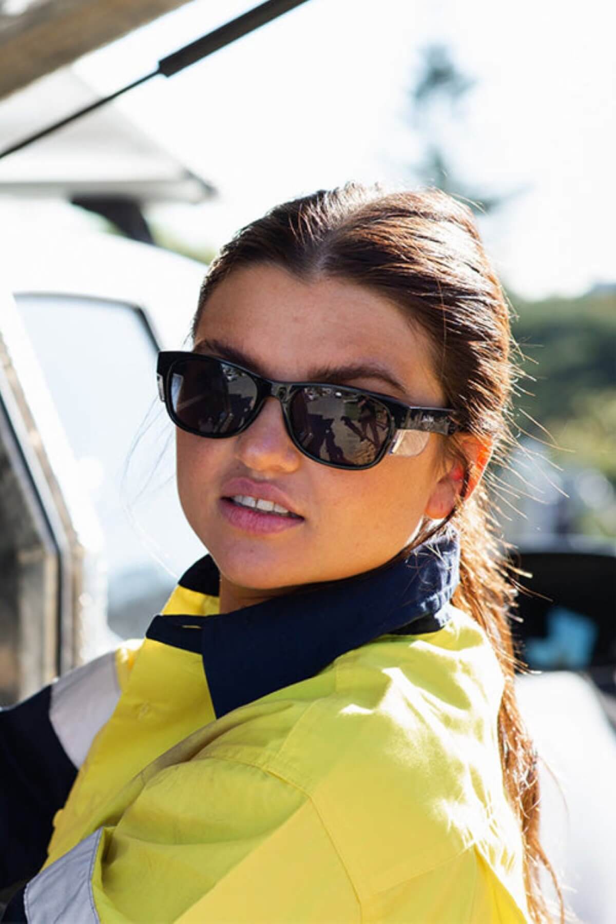A person with long hair is wearing SafeStyle Classics Black Frame Tinted Lens sunglasses and a yellow and blue high-visibility jacket. The individual is looking slightly off-camera, and the background is outdoors with some trees visible.
