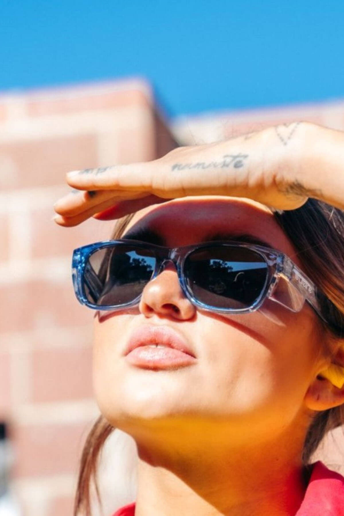 Wearing SafeStyle Classics blue frame sunglasses with polarised lenses, someone shields their eyes from the sun using their hand. A tattoo graces their wrist as they stand outdoors against a blurred brick wall and clear blue sky.