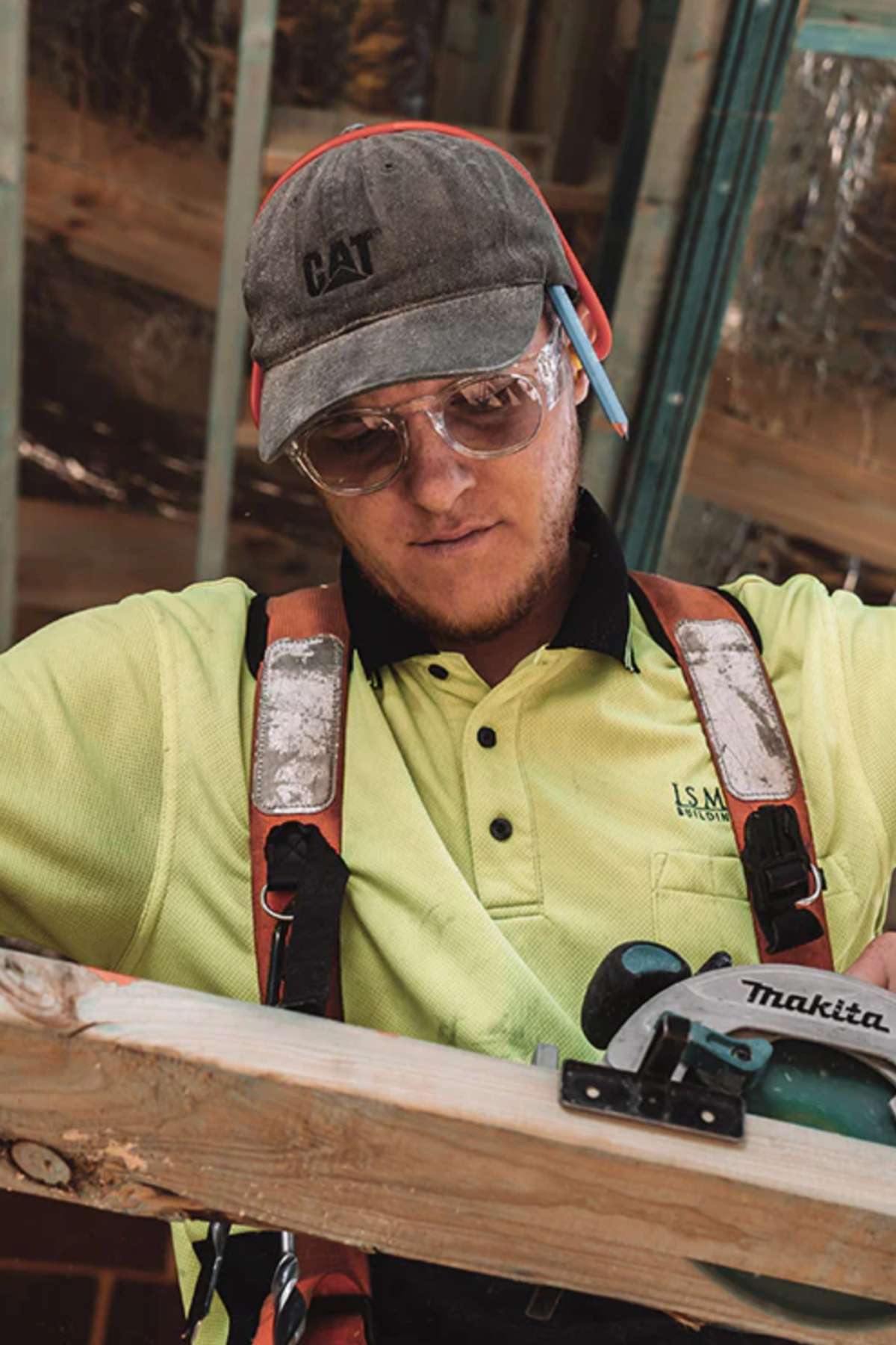 A construction worker in a yellow shirt, CAT cap, and SafeStyle Cruisers Clear Frame Hybrids Photochromic Lens safety glasses measures a wooden plank with a pencil behind his ear. A Makita saw is visible in the foreground.