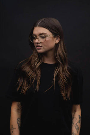 Against a dark backdrop, a person with long, wavy hair wears a black t-shirt and SafeStyle Fusions Champagne Frame Glasses. Showcasing arm tattoos, their neutral expression is visible through the lenses designed for blue light protection.
