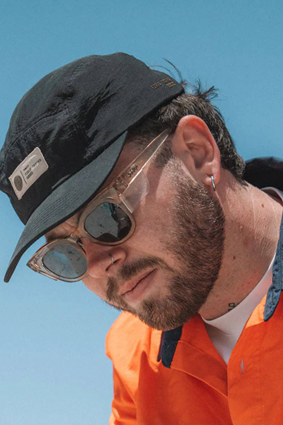A bearded man, wearing sunglasses from SafeStyle Harps with polarised lenses and an orange jacket, gazes downward thoughtfully. He sports a black cap with a small front patch and a hoop earring against a backdrop of clear blue sky.