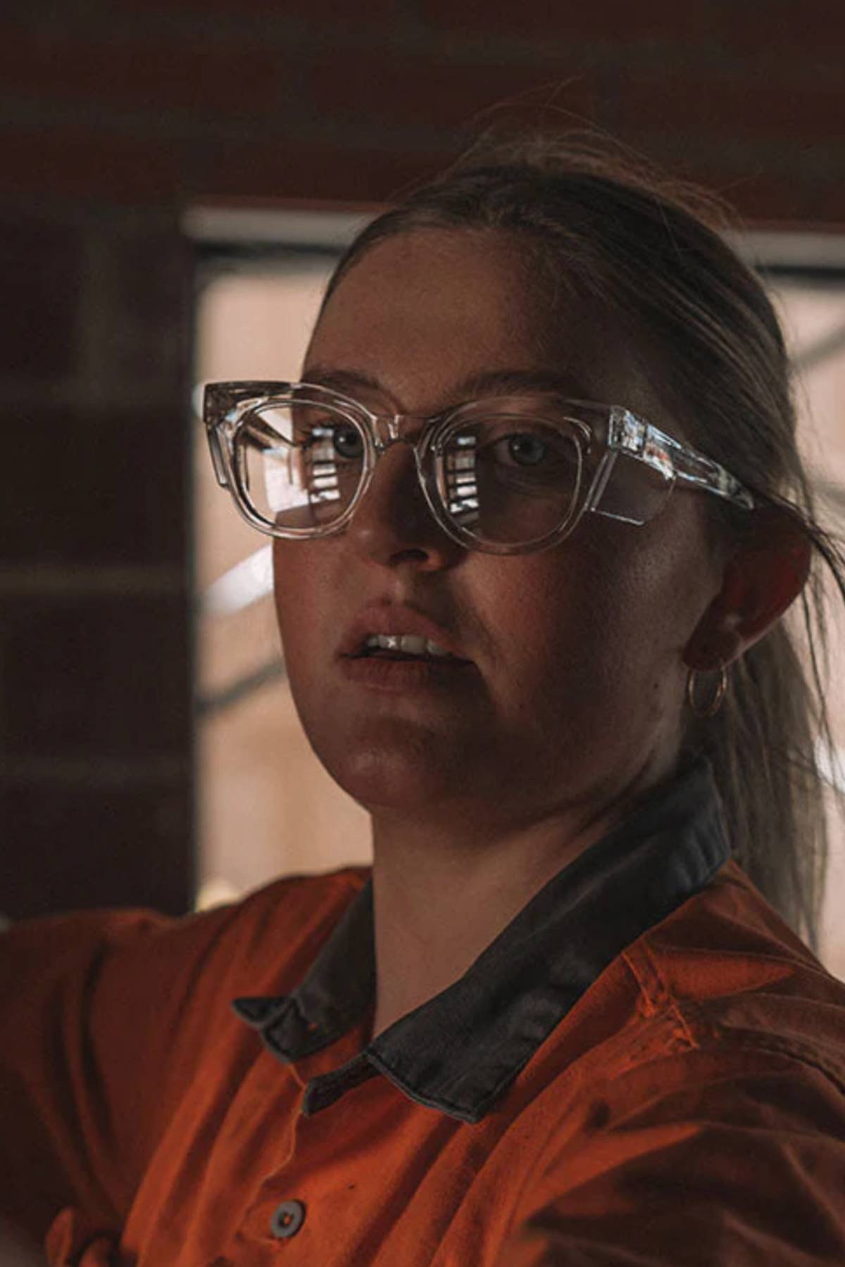 A person wearing SafeStyle Harps Clear Frame Clear Lens safety glasses and an orange shirt looks at the camera in a dimly lit room, brick wall behind them and light reflecting off their lenses.