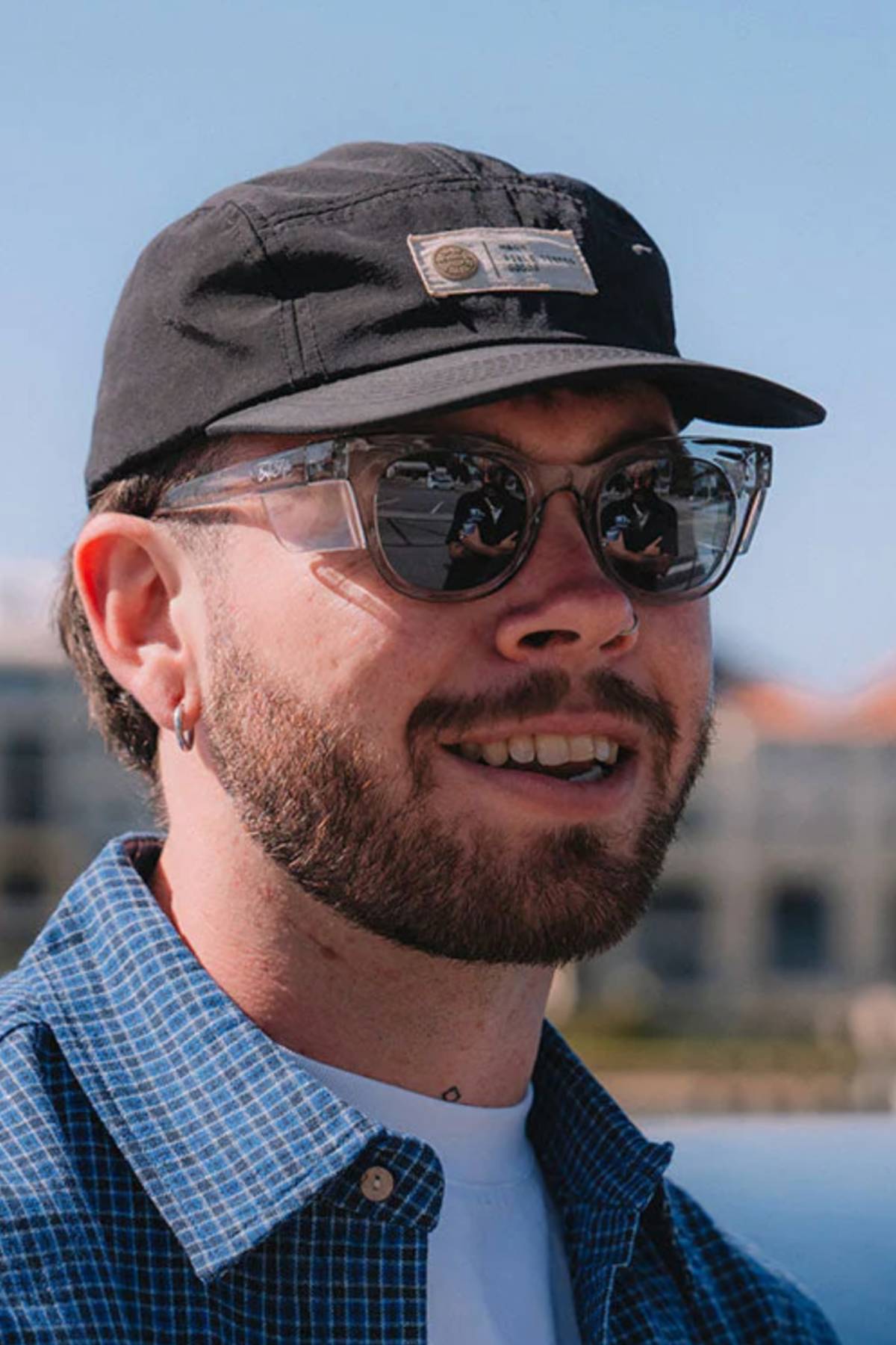 A bearded person with SafeStyle Harps graphite frame polarized sunglasses, a black cap, blue checkered shirt, and a white T-shirt smiles outdoors on a sunny day. The slightly blurred background reveals buildings bathed in sunlight.