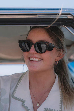 A smiling woman with long hair in a ponytail wears SafeStyle Harps Matte Tort sunglasses with brown polarised lenses. Her hoop earrings and heart-shaped necklace glimmer as she sits in a sunlit car with the window open.