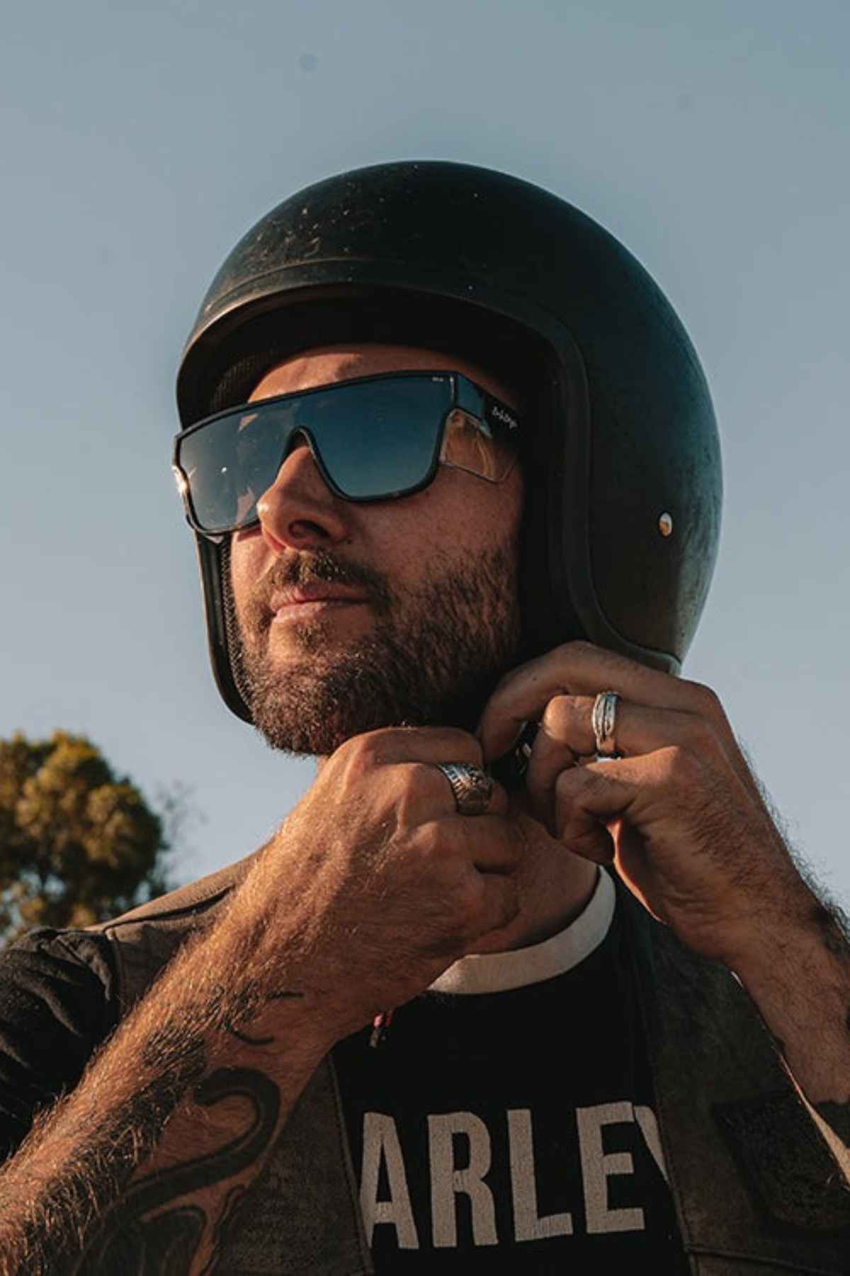 A person wearing a black helmet and SafeStyle Primes Black Frame Polarised Lens sunglasses is adjusting the chin strap of their helmet. They have a beard and are standing outdoors against a clear sky. The person is also wearing a shirt with partially visible text.