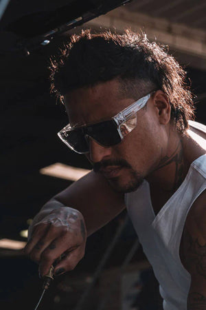 A man with a mohawk is wearing SafeStyle Primes Clear Frame Tinted Lens safety glasses featuring a shatterproof frame and antiscratch coating. In a white tank top, he's focused on a task, potentially using tools. His tattooed arms stand out in the dimly lit backdrop, adding drama to the scene.
