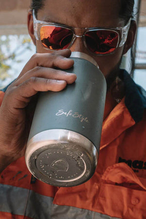 An individual in orange work attire holds a gray Safestyle Big Juicy 1L Water Bottle with double-wall vacuum insulation and a metal base while wearing reflective safety glasses. The background is blurred, emphasizing their focus on the job.