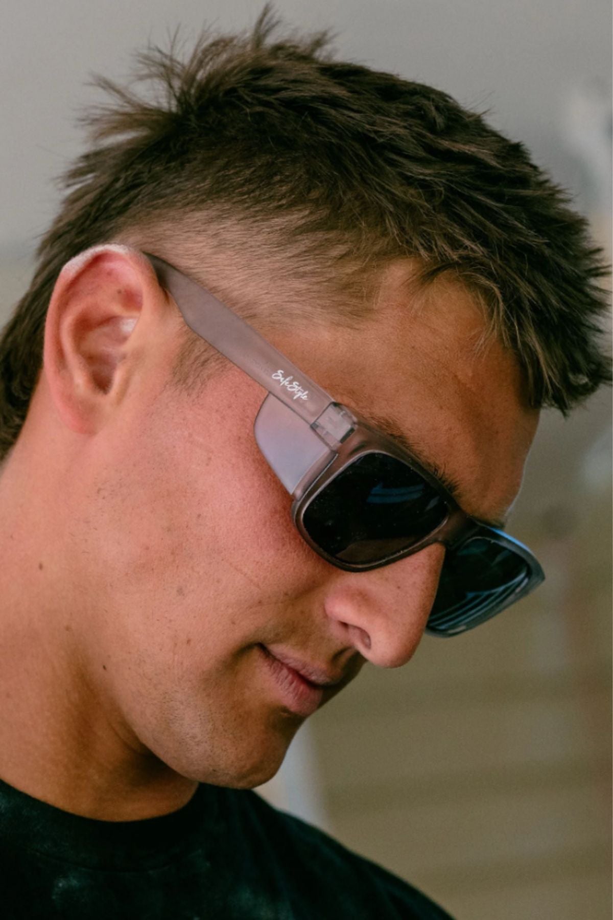 A man with short brown hair wears Safestyle Fusions XL Matte Graphite Frame Polarised Lens sunglasses and a black shirt, looking slightly downward with a neutral expression.