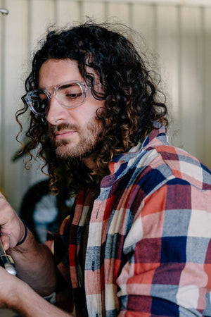 A man with long curly hair and a beard, wearing a plaid shirt and Safestyle Skylines Clear Frame Clear Lens safety glasses, is looking down and focusing on something out of frame.