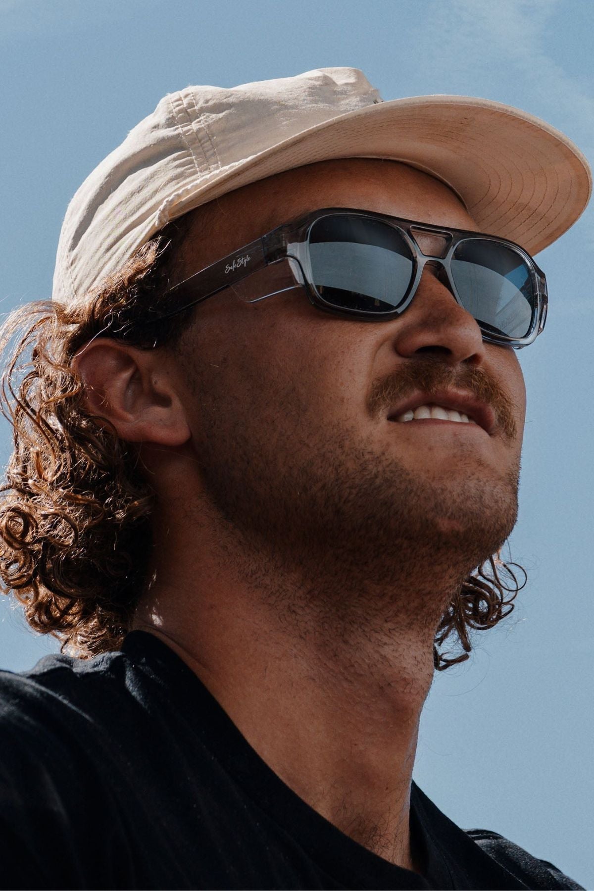 A man with curly hair in a beige cap and black shirt looks up at a clear blue sky, wearing Safestyle Skylines sunglasses with a graphite frame and grey polarised lens.