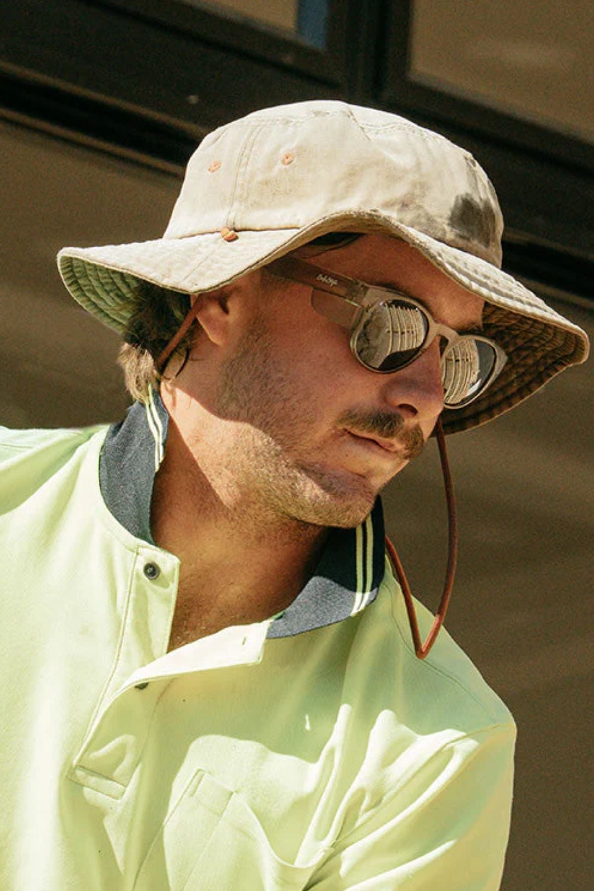 A man wearing a khaki bucket hat, Safestyle Cruisers Matte Graphite Frame Polarised Lens for UV protection, and a light yellow collared work shirt with dark trim, looking to the side in bright sunlight.