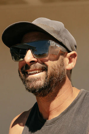 A man with a beard and mustache, wearing Safestyle Primes Matte Graphite Frame Polarised Lens sunglasses, a gray cap, and a sleeveless black shirt, smiles in bright outdoor sunlight.