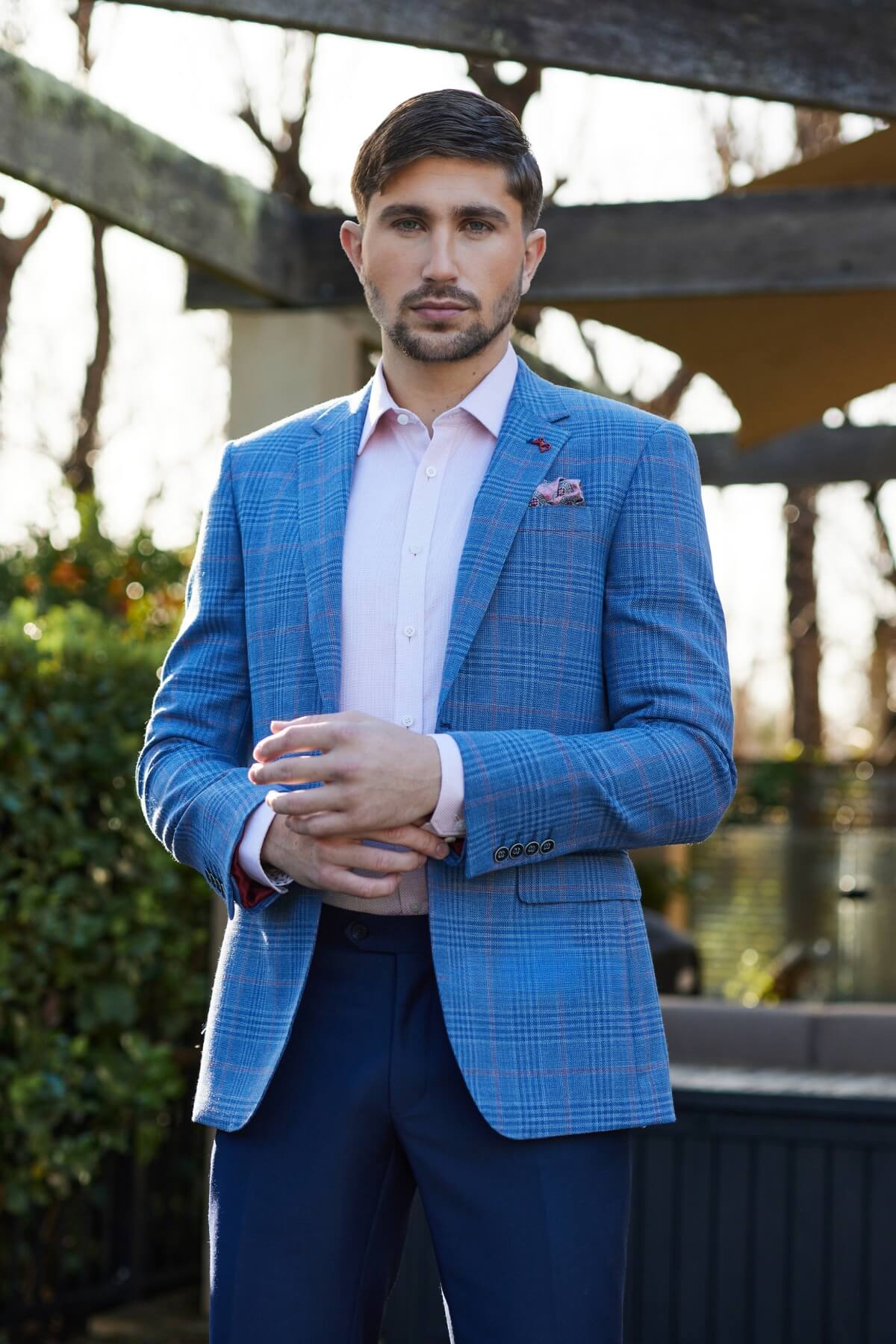 A man with short dark hair and a beard stands outdoors in the Savile Row Abram Wool Blazer, paired with a light pink dress shirt and dark blue trousers. He looks at the camera, greenery and wooden beams behind him.