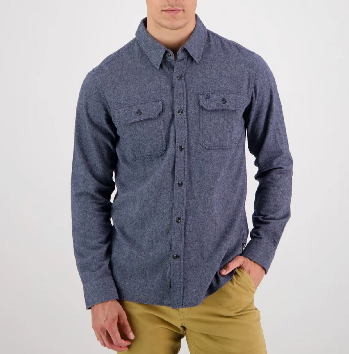 A person in a Swanndri Ranch Rd blue tailored fit shirt, 100% cotton, with two chest pockets and black buttons, pairs it with khaki pants against a white background, one hand casually in their pocket.