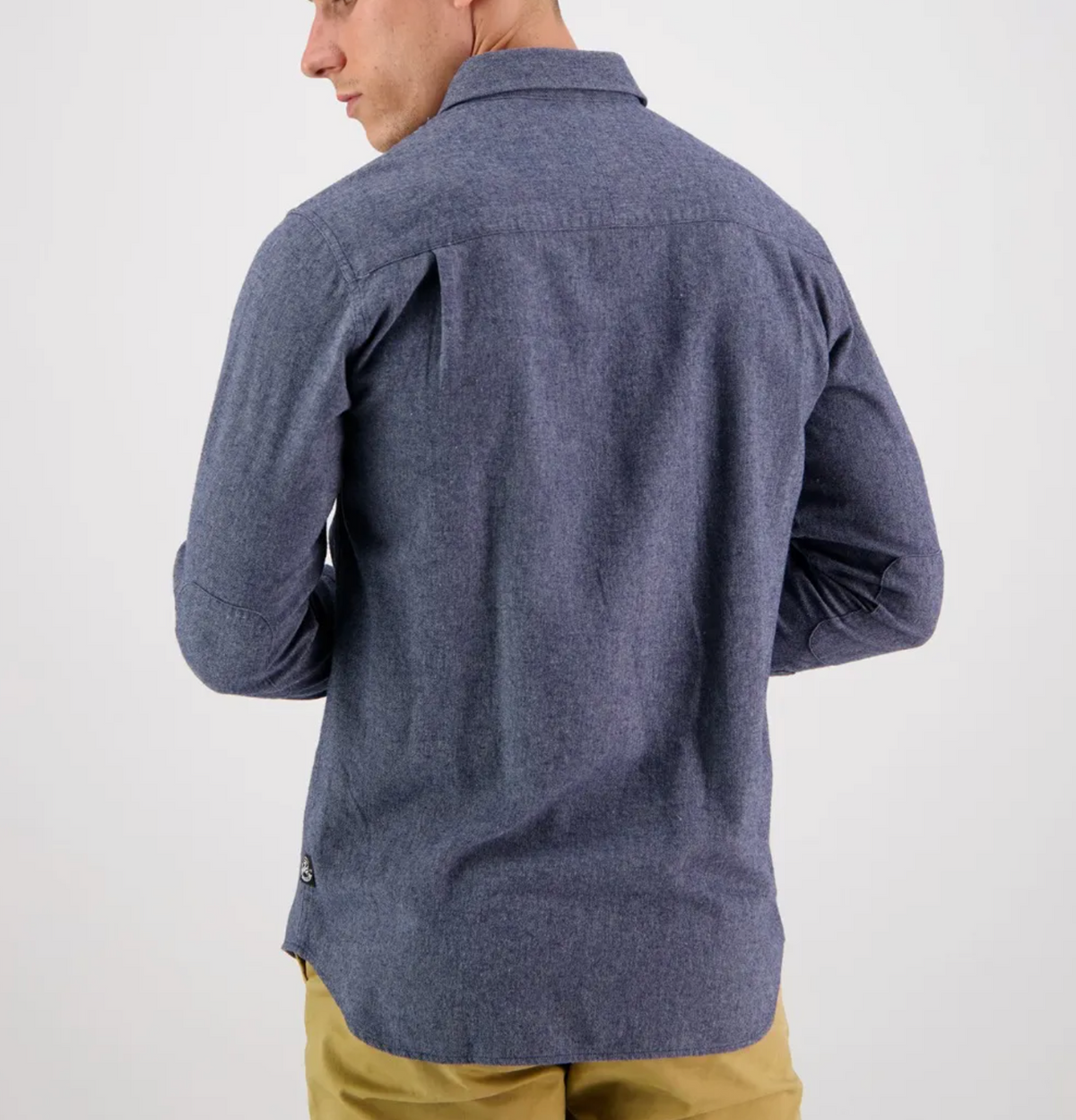 Wearing the Swanndri Ranch Rd Shirt, a 100% cotton, long-sleeved, button-up in blue, a person faces away from the camera. The perfectly tailored shirt pairs effortlessly with beige pants against a plain white background.