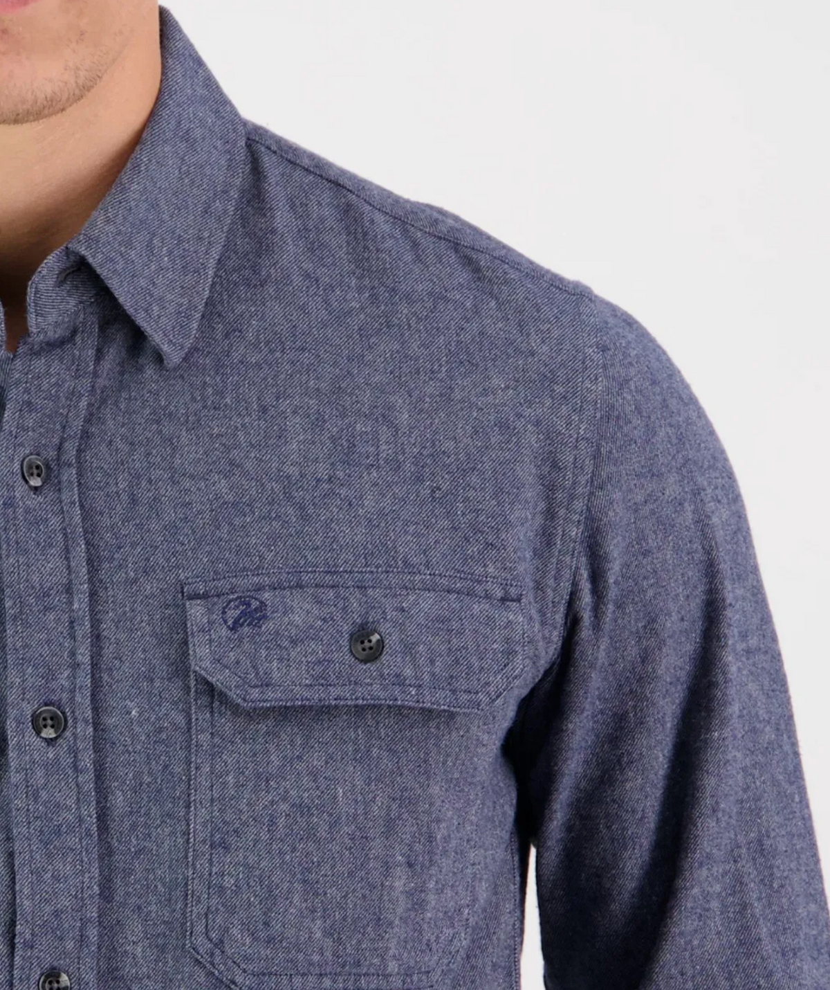 Close-up of a person wearing the Swanndri Ranch Rd Shirt in textured blue, tailored fit, and made of 100% cotton. It features a front pocket with button closure and a small embroidered design above it. The person's face is not visible against the plain white background.