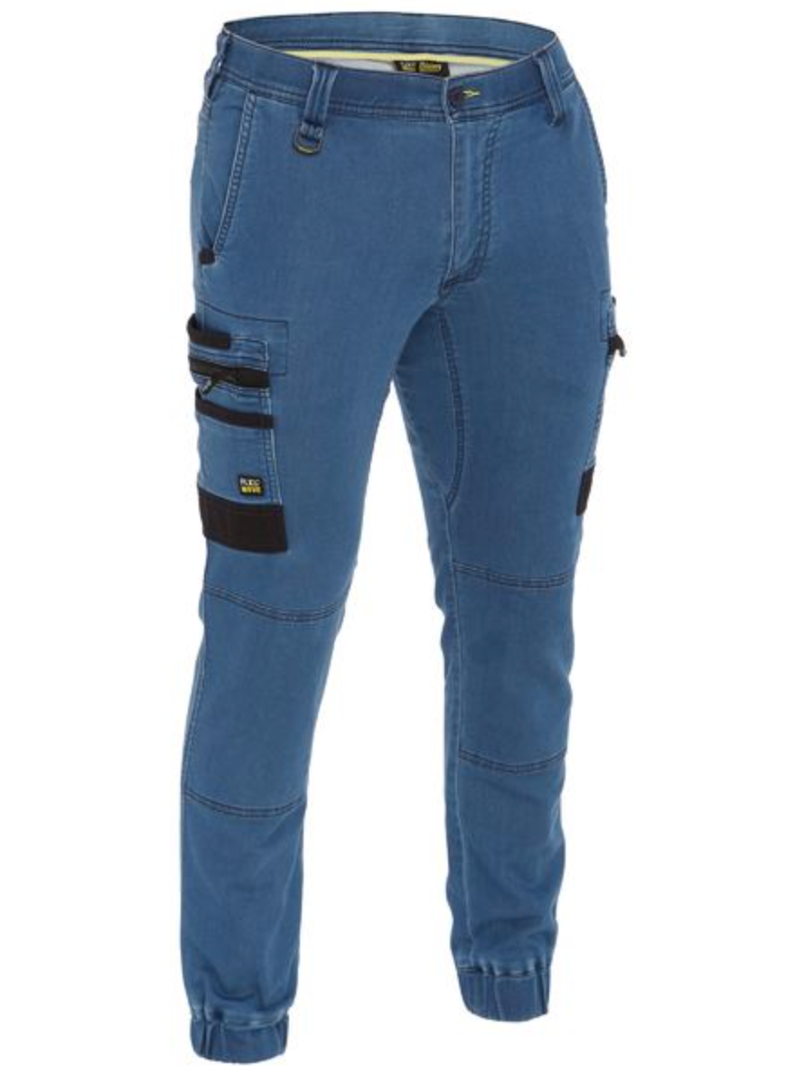 The Bisley Flex & Move Stretch Denim Cargo Cuffed Pant features blue terry knit denim for comfort, reinforced belt loops, multiple pockets with large side and small zippered options, black accents, and ribbed ankle cuffs for a modern and functional style.