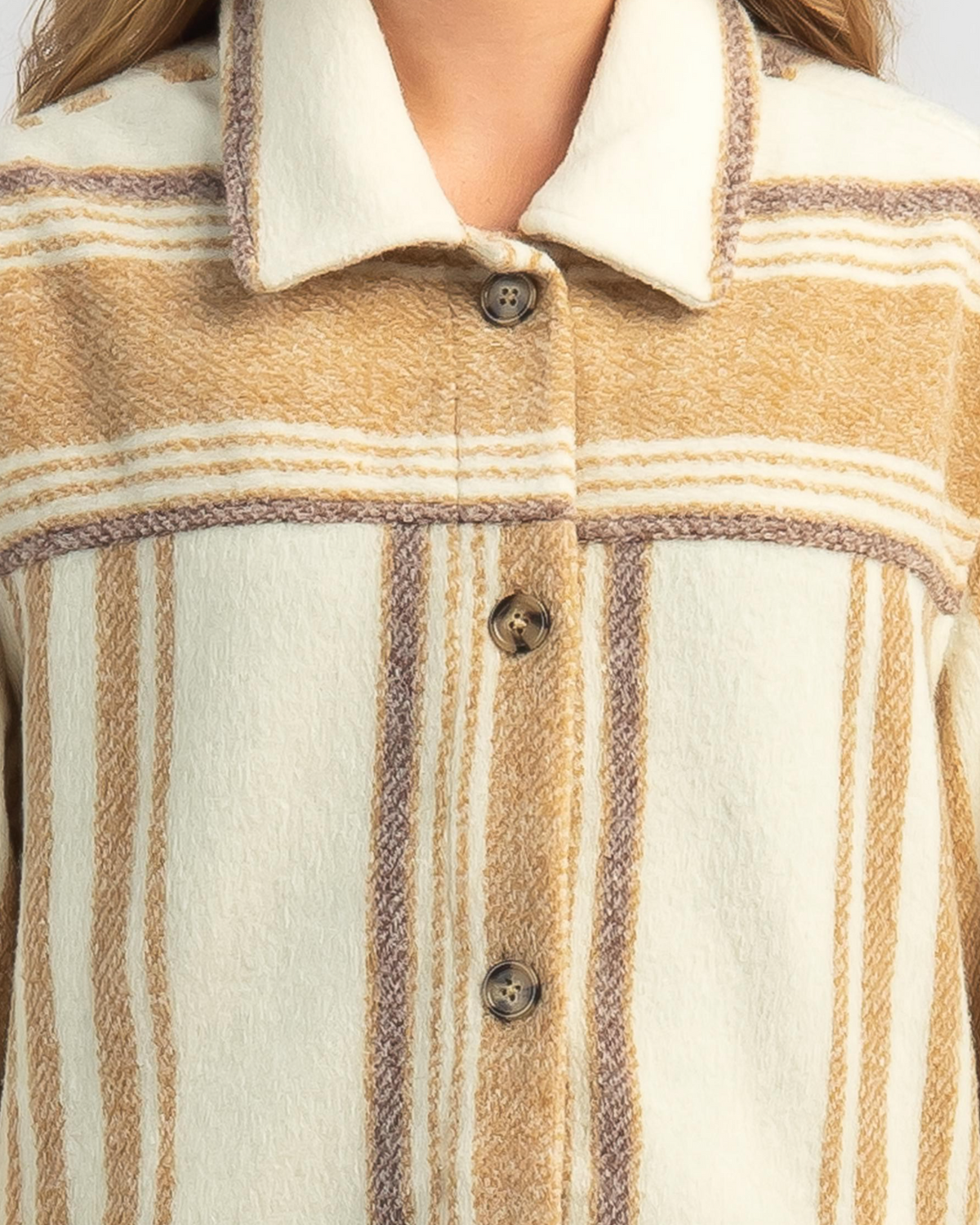 Close-up of someone in a Rip Curl Kindred Palms Jacket—a wool shacket with a cream plaid pattern, beige and brown stripes, and large front buttons. Only their lower face and upper torso are visible, exuding a casual yet stylish vibe.