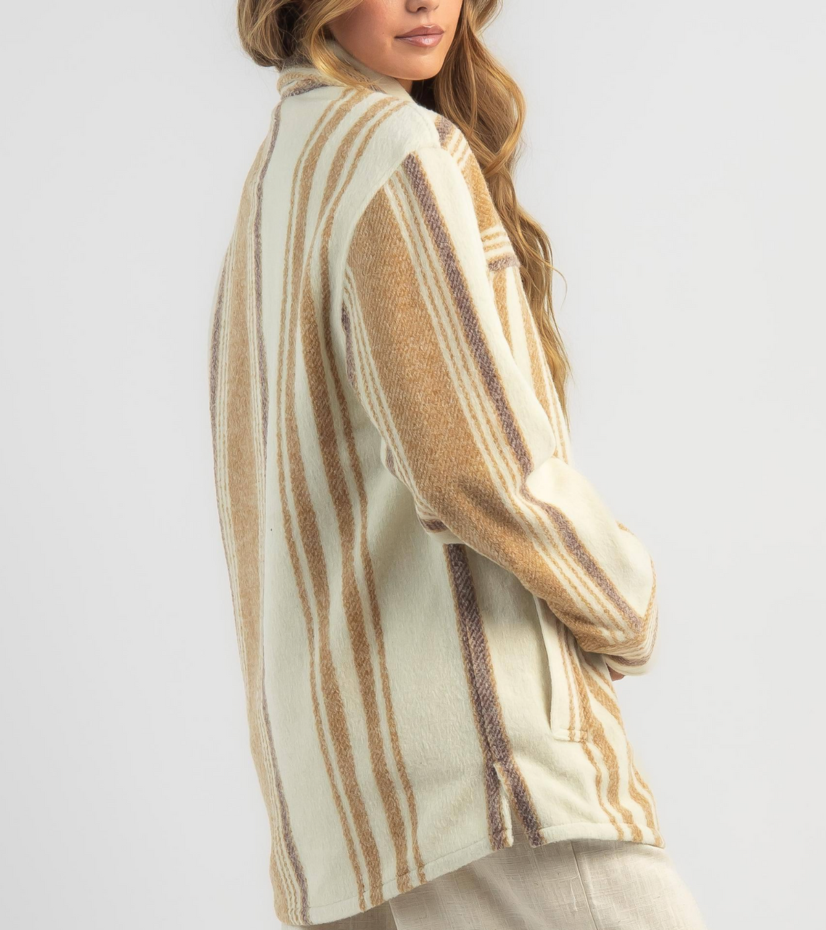 A person with long hair wears a Rip Curl Kindred Palms Jacket in cream and brown stripes, standing sideways to highlight the polyester shacket's back and side. The background is plain light gray.