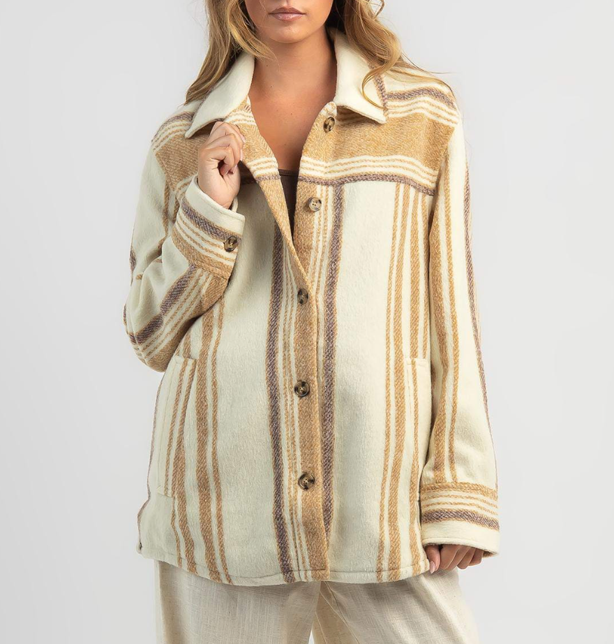 A woman wears a Rip Curl Kindred Palms Jacket in beige and brown stripes, made from soft polyester. She pairs it with light-colored pants and stands against a white background.