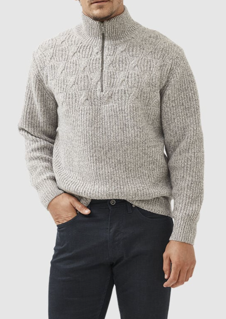 A man wearing a Rodd & Gunn Challis Knit, a gray lambswool sweater with a textured high collar and quarter zipper, pairs it with dark pants. One hand rests in his pocket while the other hangs by his side against a plain white background.