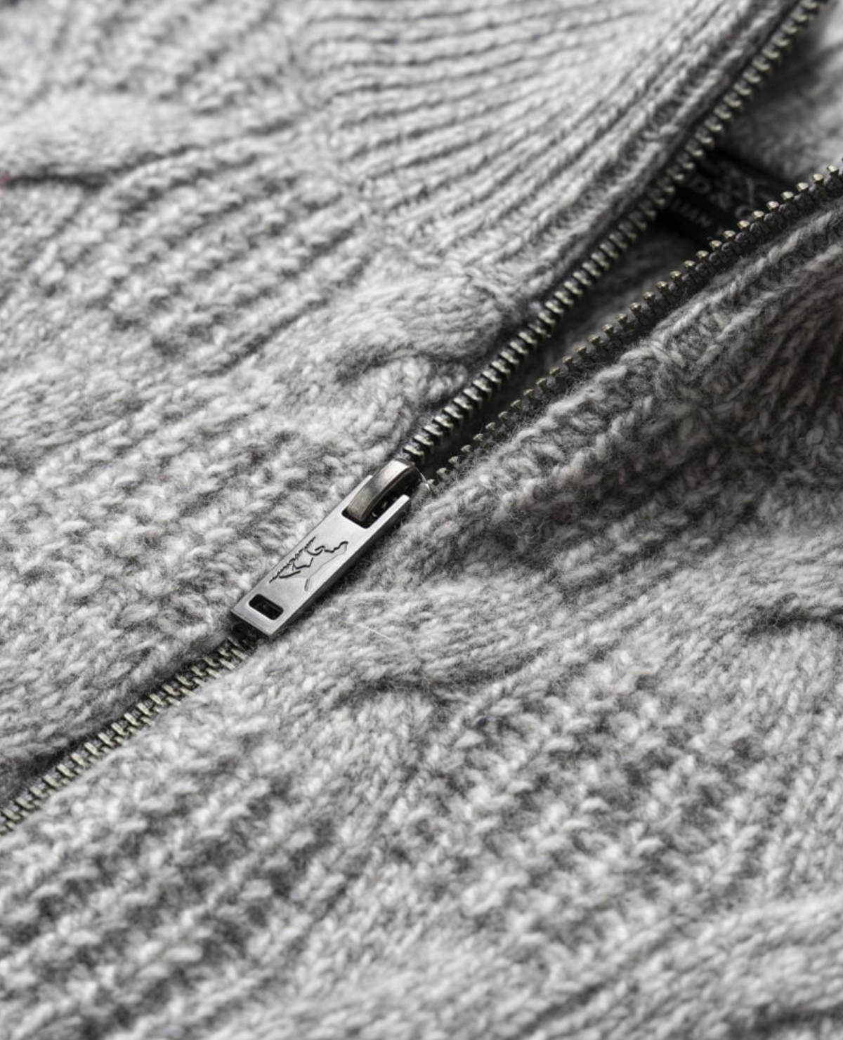 A close-up of the Rodd & Gunn Challis Knit jacket reveals its gray lambswool cable texture, featuring a partially open metal zipper that highlights the detailed fabric pattern. The zipper handle is engraved with a small logo.