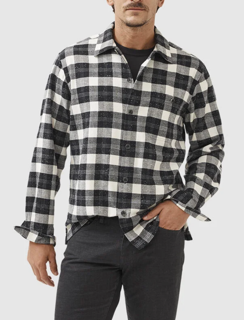 A man stands against a light background with one hand in his pocket, wearing the Rodd & Gunn Halfmoon Ave Shirt—a comfortable black and white plaid 100% cotton button-up—over a black t-shirt and black pants.