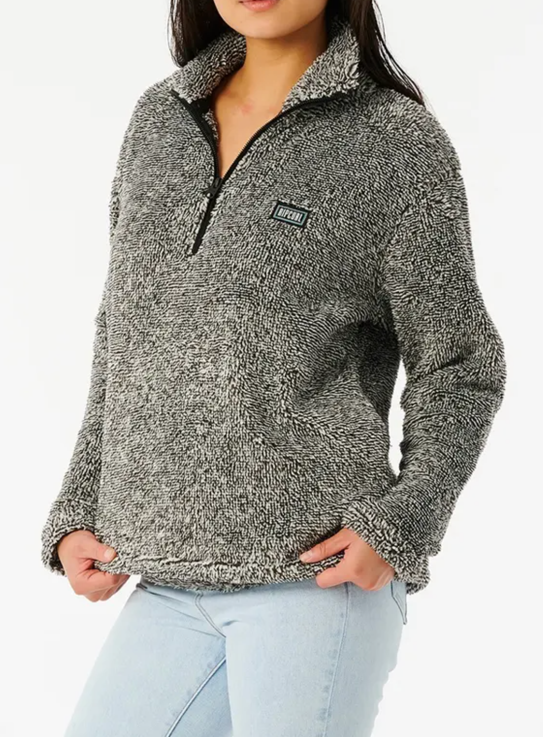 A person in a Rip Curl Dark N Stormy II cozy, textured gray half-zip pullover with a small logo on the chest has their hands in the pockets while wearing light blue jeans. The image is cropped at the neck and waist.