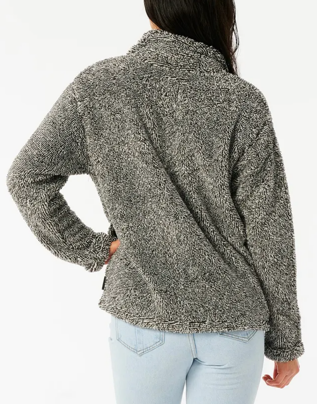 A person with long hair is wearing a cozy Rip Curl Dark N Stormy II half-zip gray sweater and light blue jeans. The person faces away from the camera, one hand on their hip, against a plain white background.