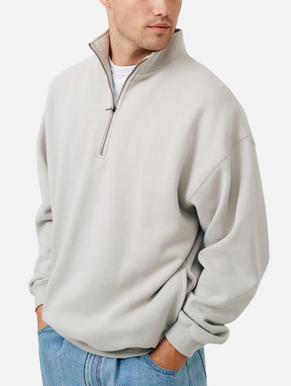 A man wears the Industrie The Del Sur Track Top in light gray with an oversized fit and soft fleece lining over a white tee and light blue jeans, standing hands-in-pockets and glancing to the side.