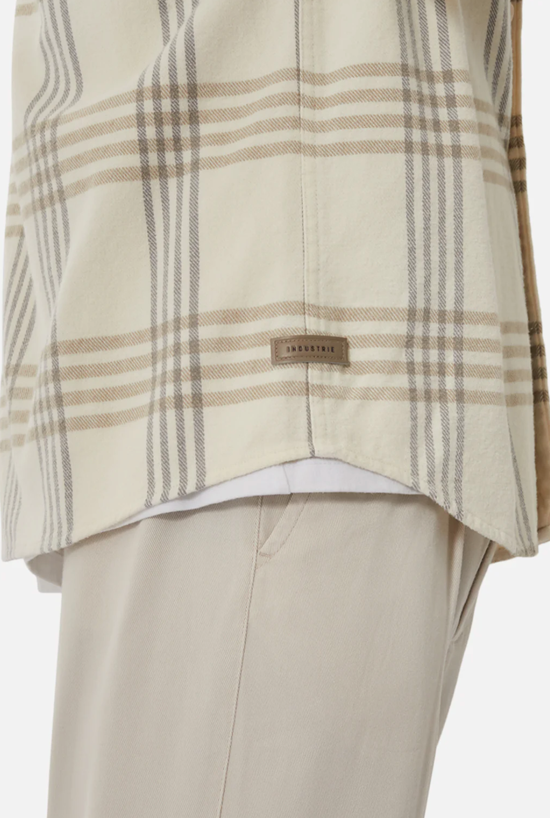 Close-up of a person wearing the Industrie The Danville Shirt, a regular fit cream plaid shirt made from 100% cotton, with tan and gray stripes over beige pants and a small "INDUSTRIE" tag near the hem.