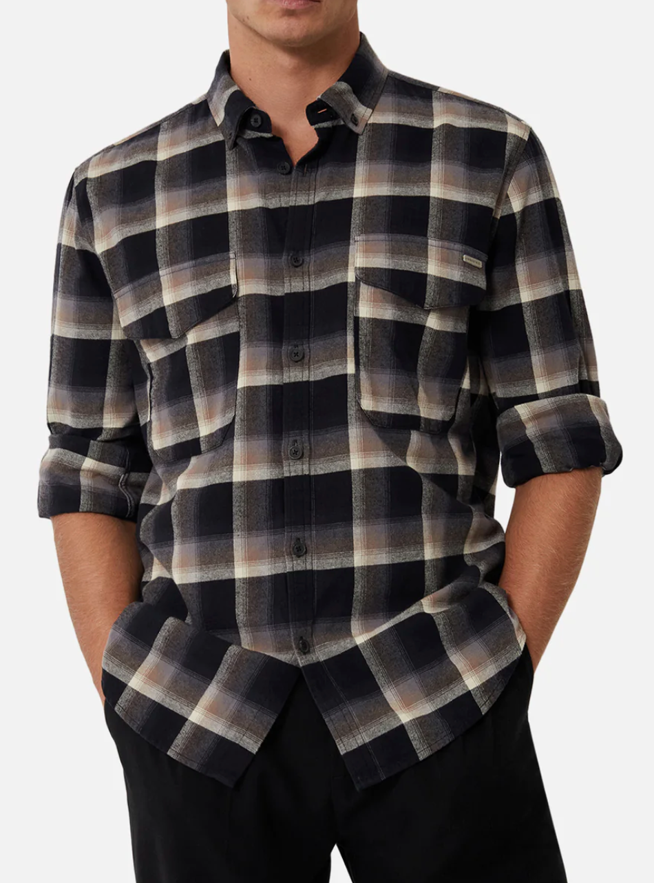 A person wearing the Industrie The Clifton Shirt, a regular fit black, brown, and beige plaid 100% cotton shirt with rolled-up sleeves, two chest pockets, and buttoned up, stands with hands in pockets. Paired with black pants, the outfit stands out against a plain white background.