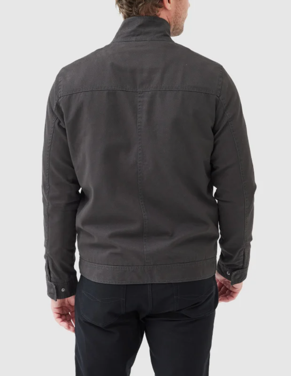 A person is standing with their back to the camera, wearing a stylish Rodd & Gunn Armitage Jacket made from Australian cotton in dark grey and black pants. The jacket features a collar and buttons on the cuffs. The background is plain light grey.