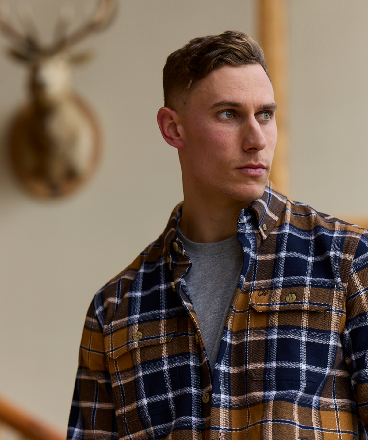 A man in a Swanndri Pencarrow Shacket, classic fit and 100% cotton plaid, layered over a gray t-shirt looks to the side with a blurred deer head mounted on a beige wall in the background.