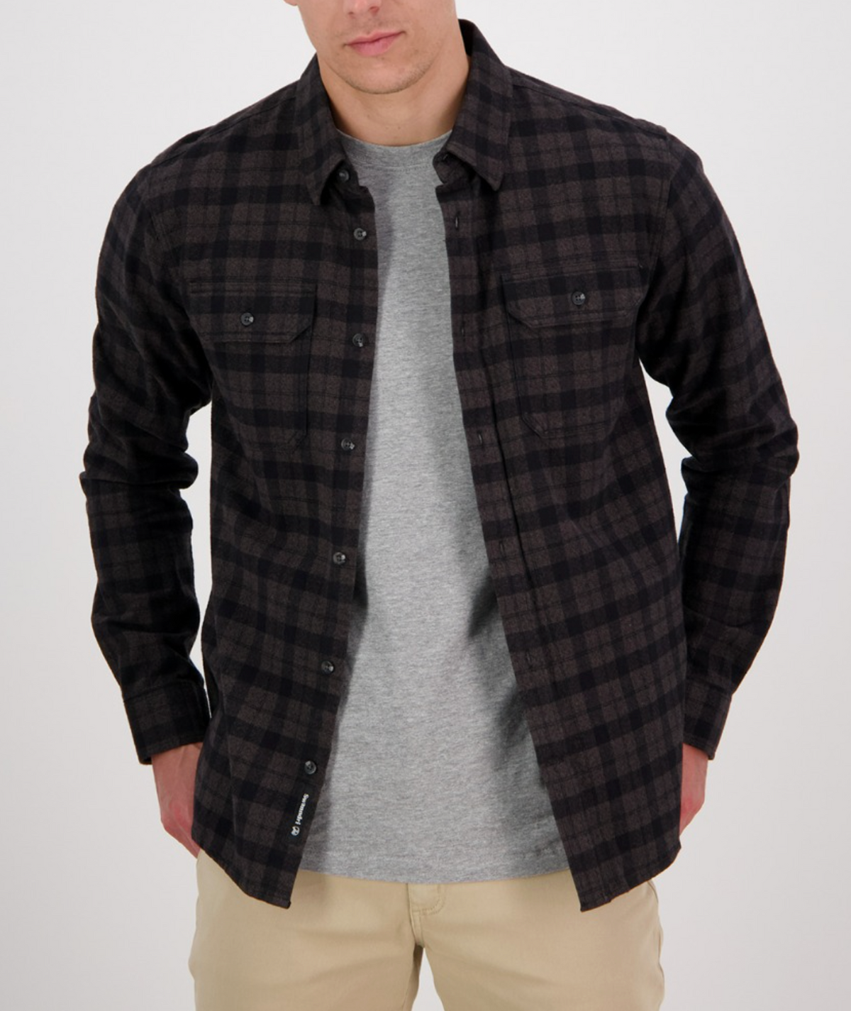 A person wears a Swanndri Port Road Shirt, featuring a dark plaid classic twill design, unbuttoned over a gray T-shirt with beige pants. The 100% cotton shirt complements the look as they stand with hands in pockets against a plain background, face not visible.